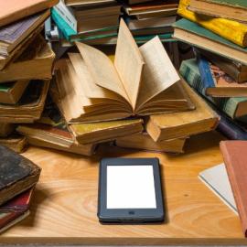Books stacked haphazardly, an eReader placed on the table in front of the books